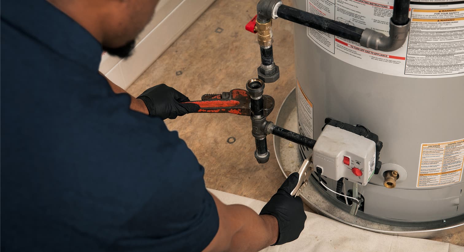A Maitz plumber wearing black gloves uses a pipe wrench to repair the gas line and control valve assembly on a residential water heater tank.