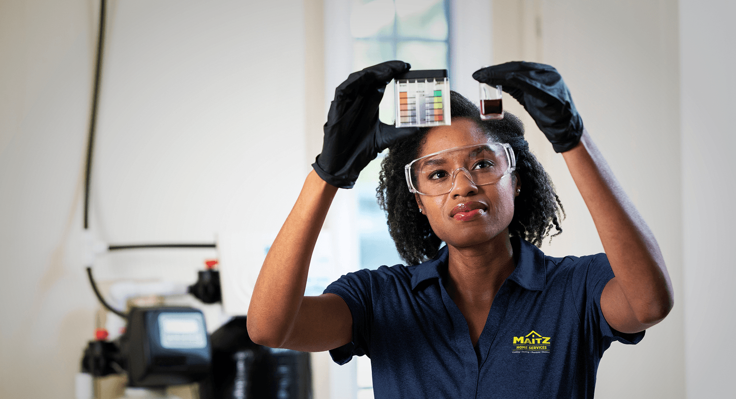 A Maitz technician in a blue uniform and safety gear compares a water sample to a testing chart in front of a water softener system.